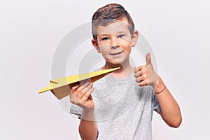 Cute blond kid holding paper airplane smiling happy and positive, thumb up doing excellent and approval sign