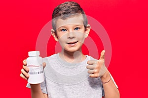 Cute blond kid holding pills smiling happy and positive, thumb up doing excellent and approval sign