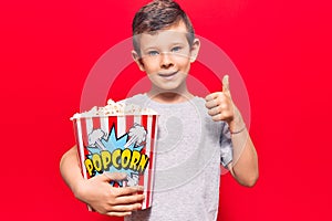 Cute blond kid holding popcorn smiling happy and positive, thumb up doing excellent and approval sign