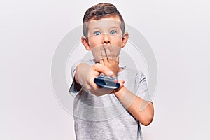Cute blond kid holding television remote control covering mouth with hand, shocked and afraid for mistake