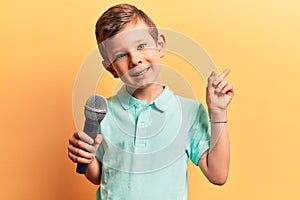 Cute blond kid singing song using microphone smiling happy pointing with hand and finger to the side