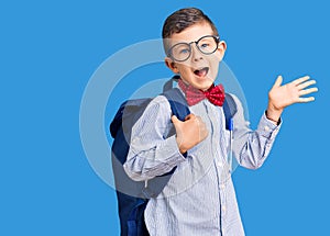 Cute blond kid wearing nerd bow tie and backpack celebrating victory with happy smile and winner expression with raised hands