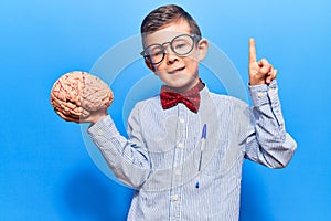 Cute blond kid wearing nerd bow tie and glasses holding brain smiling with an idea or question pointing finger with happy face,