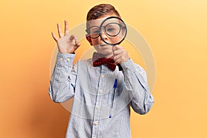 Cute blond kid wearing nerd bow tie and glasses holding magnifying glass doing ok sign with fingers, smiling friendly gesturing