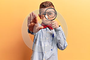 Cute blond kid wearing nerd bow tie and glasses holding magnifying glass with open hand doing stop sign with serious and confident