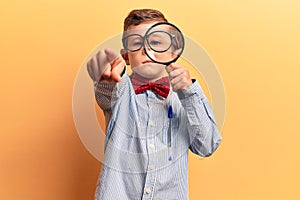 Cute blond kid wearing nerd bow tie and glasses holding magnifying glass pointing with finger to the camera and to you, confident