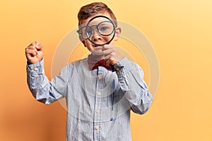 Cute blond kid wearing nerd bow tie and glasses holding magnifying glass screaming proud, celebrating victory and success very