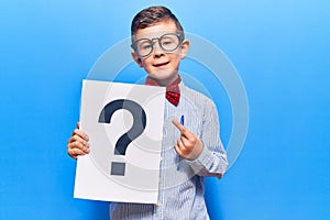 Cute blond kid wearing nerd bow tie and glasses holding question mark smiling happy pointing with hand and finger