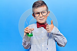 Cute blond kid wearing scientist bow tie and glasses holding test tube smiling happy and positive, thumb up doing excellent and