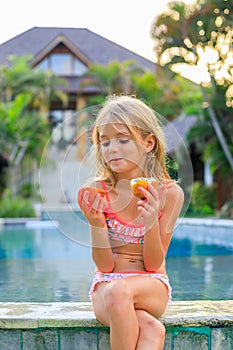 Cute blond little girl in pink swimsuit sitting at the swimming pool, holding passion fruit. Enjoy eating tropical fruit. Summer photo