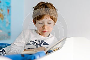 Cute blond little kid boy in pajamas reading book in his bedroom. Excited child reading loud, sitting in his bed