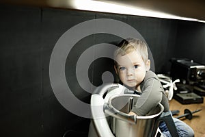 Cute blond little toddler boy mix food with hand in steel blender, food processor, sitting on kitchen drawer. Copy space