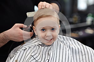 Cute blond smiling baby boy with blue eyes in a barber shop having haircut by hairdresser. Hands of stylist with tools. Children`s