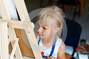 Cute blond smiling caucasian kid painting on wooden easel in class workshop lesson at art studio. Little girl holding brush in