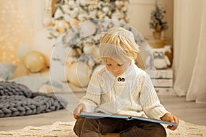Cute blond toddler preschool boy, reading a book and opening present on Christmas on cozy home