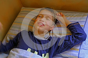 Cute blonde boy in blue pajama smiling, waking up in bed in the morning close-up. Kids bedroom. Relax