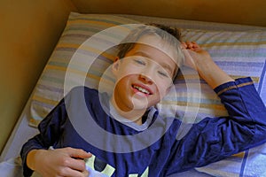 Cute blonde boy in blue pajama smiling, waking up in bed in the morning close-up. Kids bedroom. Relax