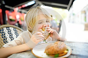 Cute blonde boy eating large bagel with salmon and arugula in outdoor fast food restaurant