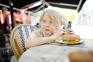 Cute blonde boy eating large bagel with salmon and arugula in outdoor fast food restaurant