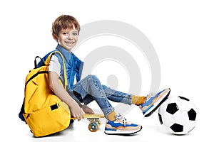 Cute blonde boy sitting on yellow skateboard and holding leg on soccer ball on white background. Back to School.