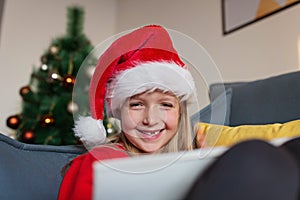 Cute blonde caucasian girl 7 years old sitting on couch near Christmas tree and writing letter to Santa Claus. Bedroom