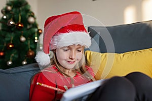 Cute blonde caucasian girl 7 years old sitting on couch near Christmas tree and writing letter to Santa Claus. Bedroom