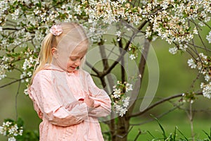Cute blonde girl in blooming garden hugging herself. Little princess in pink dress stands near flowering tree