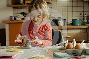 Cute blonde girl decorating Easter eggs at home.