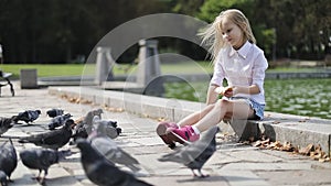 Cute blonde girl feeding pigeons bread in the park near the lake.