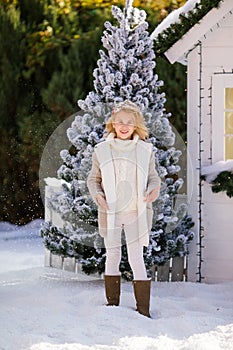 Cute blonde girl under the falling snow near the small house and snow-covered trees. New Year and Christmas time