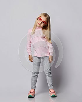 Cute blonde kid girl in stylish smoked heart-shaped sunglasses, turtleneck shirt sweater and pants is posing over gray