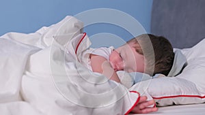 Cute blonde little boy sleeps alone at home in bed with teddy bear in snow-white clean bed. Boy dreams, sleeps badly