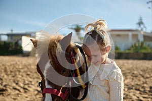 Cute blonde little child girl stroking a pony, smiling and enjoying happy weekend outdoor. Children and love for animals