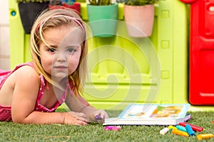 A cute blonde little girl laying on the grass and colouring a book.