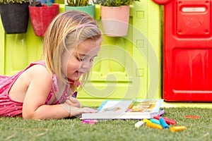 A cute blonde little girl laying on the grass and colouring a book.