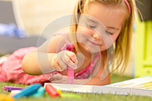 A cute blonde little girl laying on the grass and colouring a book.