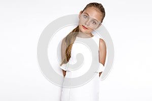 Cute blonde little girl in a white elegant dress, standing on a white background in studio.