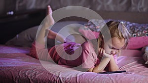 Cute blonde small girl dressed in pink pyjamas is lying in bed and watching cartoons with smartphone. Toddler female kid