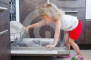 Cute blonde toddler girl helping in the kitchen taking plates out of dish washing machine