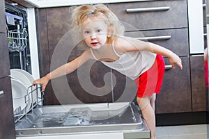 Cute blonde toddler girl helping in the kitchen taking plates out of dish washing machine