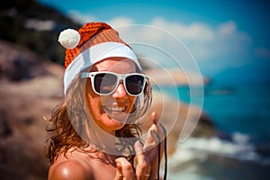 Cute blonde woman in bikini, sunglasses and santa hat on at exotic tropical beach. Holiday concept for New Years Cards.