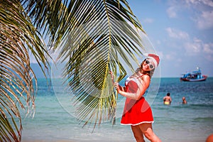 Cute blonde woman in red dress, sunglasses and santa hat stands at palm tree on exotic tropical beach. Holiday concept