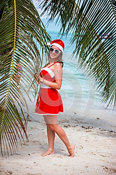 Cute blonde woman in red dress, sunglasses and santa hat stands at palm tree on exotic tropical beach. Holiday concept