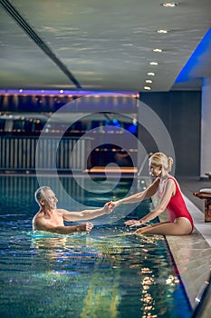 Cute blonde woman in red swimming suit giving hand to her husband in na swimming pool