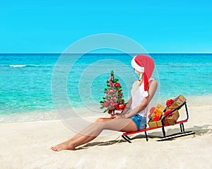 Cute blonde woman at sea beach in santa hat sitting at sled with