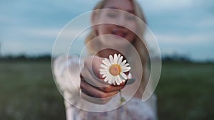 Cute blonde young woman showing a daisy to the camera standing on the field