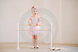 A cute blue-eyed baby ballerina in a pink leotard and tutu standing near a ballet barre. Dream concept of becoming a ballerina
