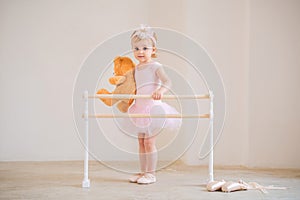 Cute blue-eyed baby ballerina in pink, standing near the ballet barre with a teddy bear in her hands. Dream concept of becoming a