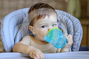 Cute blue-eyed baby boy holding plastic mug in his hands and drinking water itself lookin at camera,