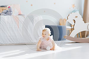 A cute blue eyed blonde one year old baby in a pink dress is holding crawling on all fours in the children`s room on the floor